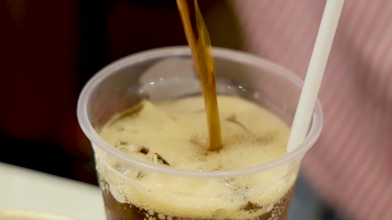 A hand pours dark soda from a can into a clear plastic cup filled with ice and a straw, creating foam. Indoor, close-up, steady lighting