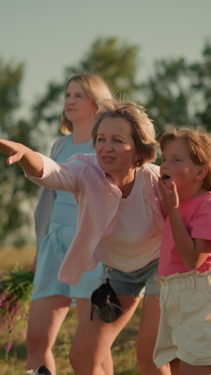 다른 여자가 꽃을 들고 배경에 서있는 동안 어머니는 그녀의 딸에게 멀리서 흥미로운 것을 지적하면서 언덕의 농장에서 서있는 가족