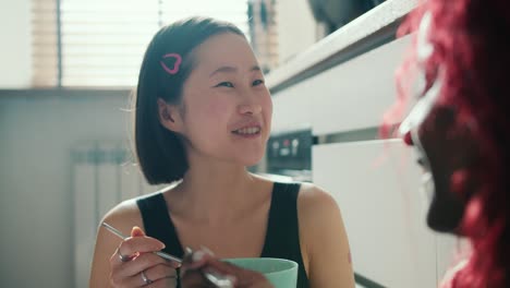 asian girl eats breakfast from a bowl with her friend sitting on the floor in the kitchen and talk