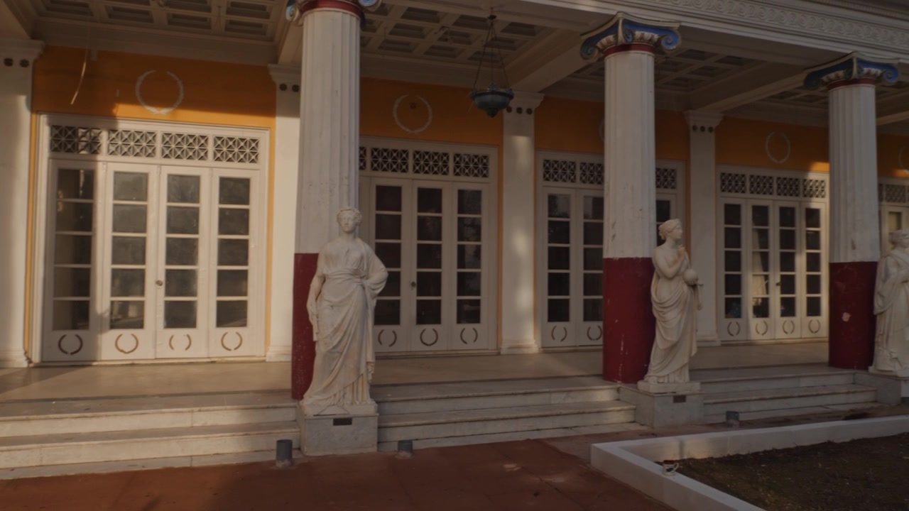 Langsom kamerapanorering langs Musenes Peristyl ved Achilleion-palasset på Korfu, Hellas. Klassiske marmorstatuer og elegante søyler bader i varmt sollys