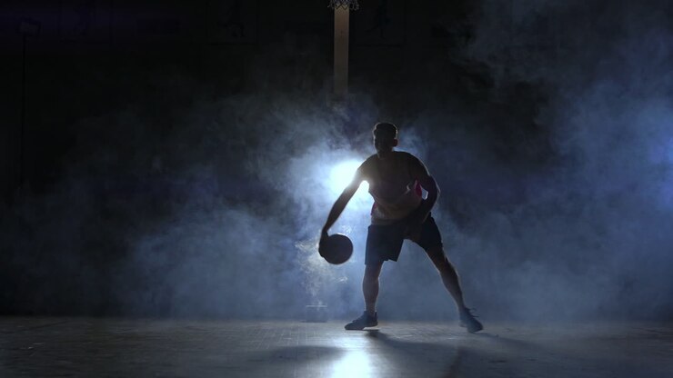 A man with a basketball on a dark basketball court against the backdrop of a basketball ring in the smoke shows dribbling skills illuminated by three lanterns in backlight