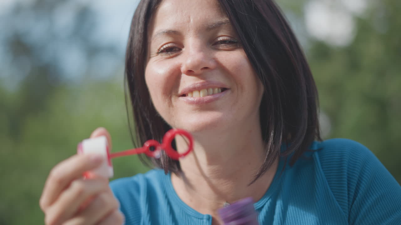 Close-up de uma mulher caucasiana soprando bolhas de sabão, retrato em um parque ensolarado com uma expressão alegre, anéis de bolhas flutuando em direção à câmera, risadas espontâneas e sorrisos largos retratando uma mãe brincalhona em um piquenique