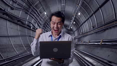 asian male professional worker standing with his laptop in underground subway tunnel, he raises his fist up with screaming goal