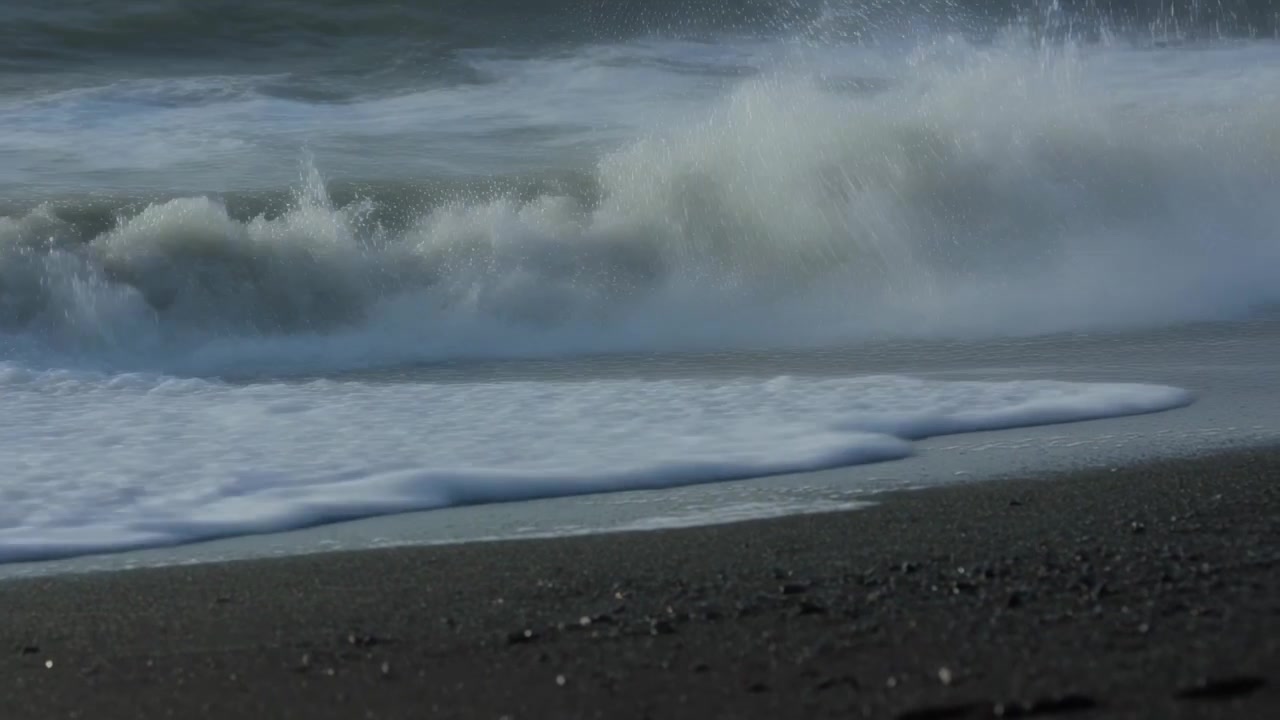 Moving swell pushing breaking ocean waves onto low-angle wet dark pebbly beach, creating white foam