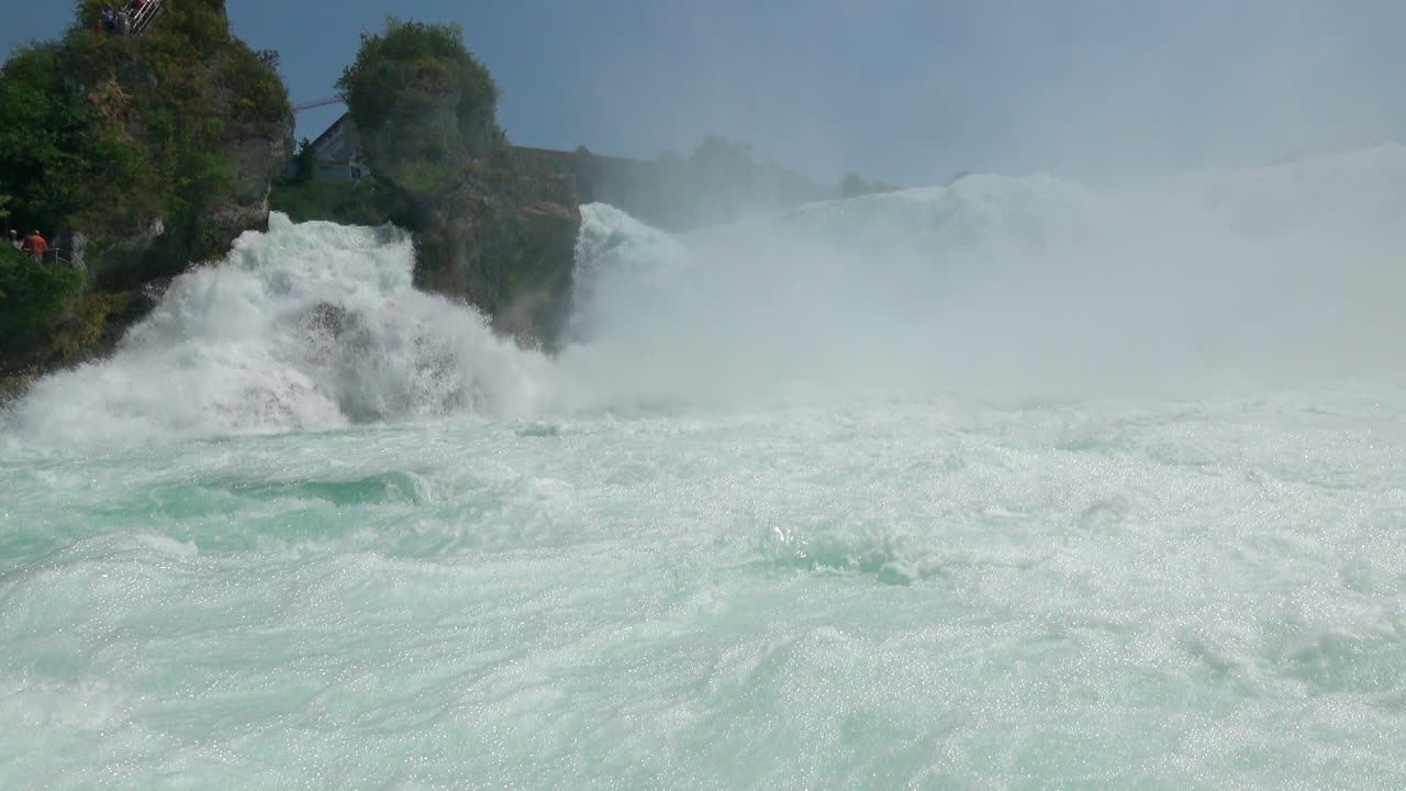 Wide view of Rhine Falls with powerful cascading water, spray and turquoise river surface, surrounded by rocky cliffs and trees near Schaffhausen, Switzerland
