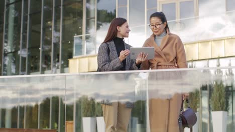 Two-Young-Successful-Intercultural-Businesswomen-Looking-Through-Online-Goods-On-Touchpad-Display-While-Standing-By-Office-Center-After-Work