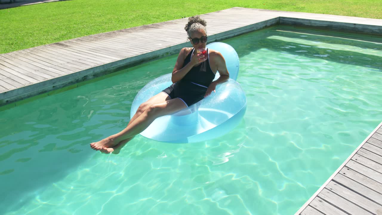 African American woman lounging on blue float in backyard pool in black swimsuit, sipping red cup