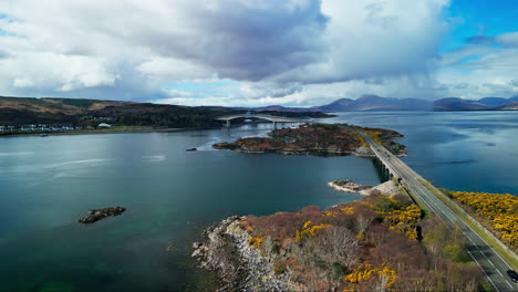 drone rises above rocky shoreline and yellow fields above islands and skye bridge in scotland