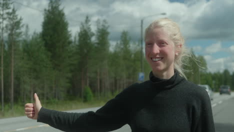 hermosa chica rubia que hace autostop en un cruce de carreteras, campo finlandés, mujer que hace autostop con una mochila pesada en escandinavia, los coches no se detienen, primer plano en la cara