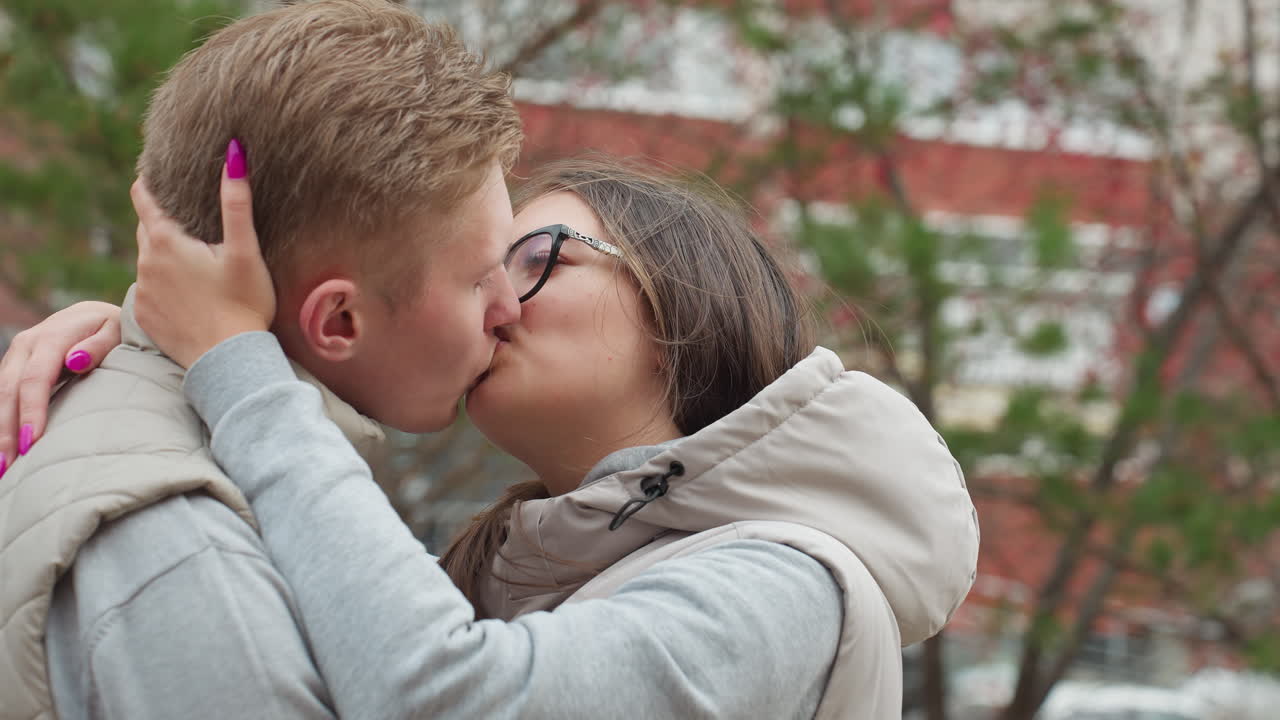 Two lovers share passionate kiss outdoors in matching outfits, wind gently sways hair and autumn leaves around them as they embrace closely