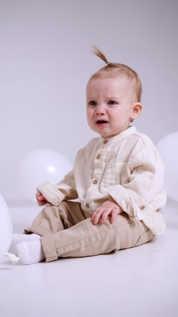 Little Caucasian baby in light clothes is starting to cry. Weepy toddler child sitting on the floor. White backdrop. Vertical video.