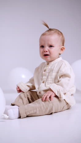 little caucasian baby in light clothes is starting to cry. weepy toddler child sitting on the floor. white backdrop. vertical video.