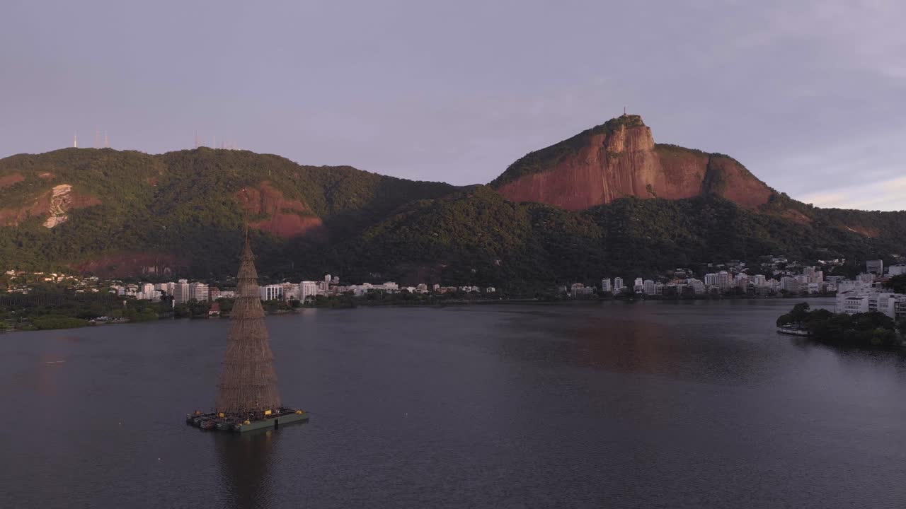 movimento panorâmico mostrando o lago da cidade do rio de janeiro com no meio a árvore de natal flutuante mais alta de 2018 com ao fundo o morro do corcovado com a estátua do cristo ao nascer do sol