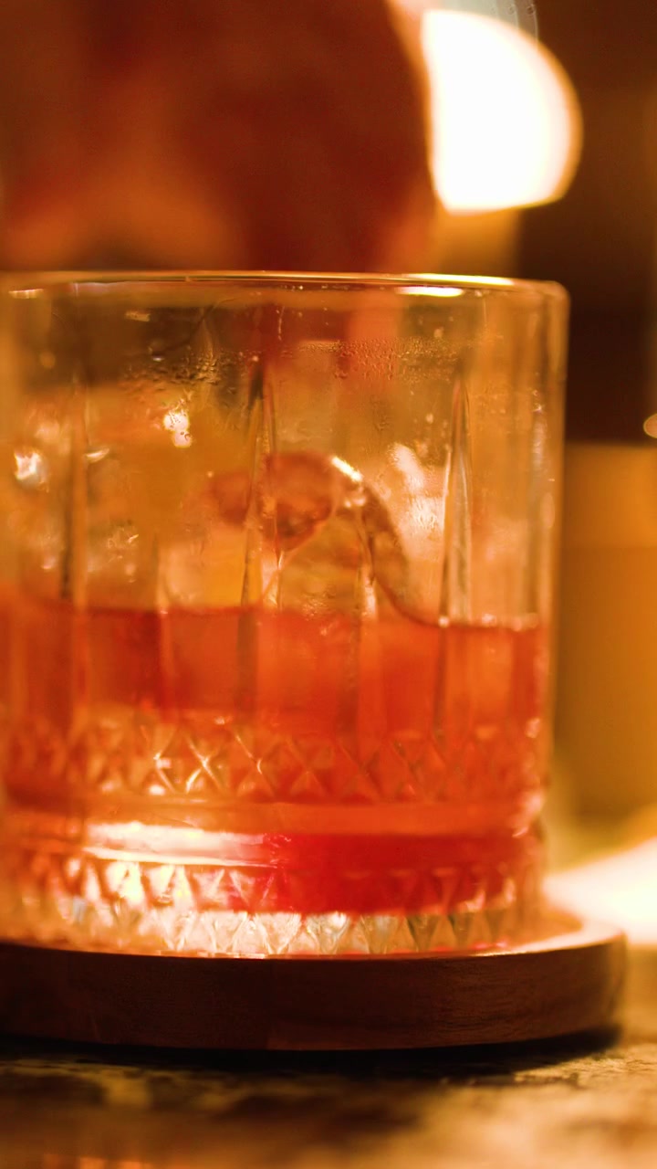 Una mano solleva un bicchiere da cocktail con ghiaccio da un bancone del bar sotto un'illuminazione ambrata