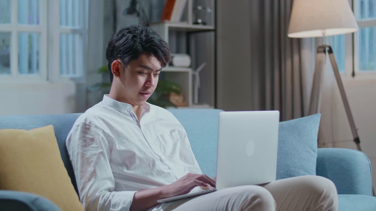 Smiling Asian Man Typing On Laptop Computer While Sitting On Sofa In The Living Room