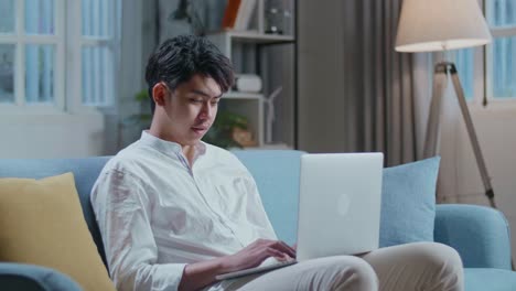 smiling asian man typing on laptop computer while sitting on sofa in the living room