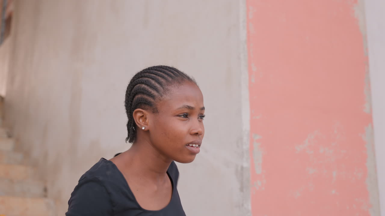 Donna di colore che parla accanto a un muro rosa. Acconciatura con treccine aderenti, profilo spontaneo, bocca espressiva, gesto del braccio esteso, pianerottolo di scala esterno, narrazione emotiva, tono colloquiale, scena urbana intima.