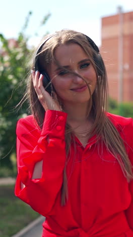 beautiful young woman with headphones. happy girl walking and enjoying listening to music in the street. attractive female in red dress on the city background. vertical video