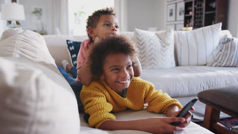 Niña-Preadolescente-Tumbada-En-El-Sofá-Viendo-La-Televisión,-Su-Hermano-Menor-Sentado-Sobre-Su-Espalda,-De-Longitud-Completa