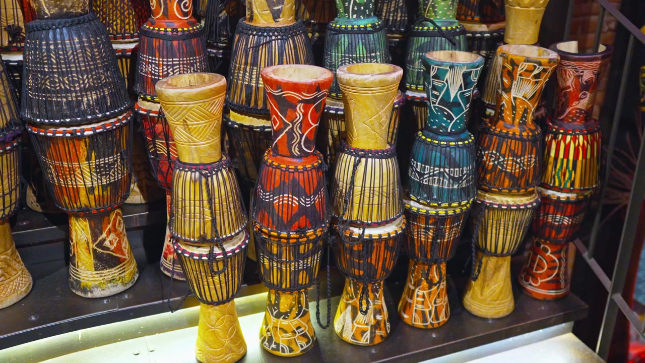 Una vivace collezione di tamburi djembe tradizionali, ognuno dipinto a mano con audaci motivi tribali africani nei toni del rosso, blu, giallo e arancione, esposti insieme su uno scaffale del mercato