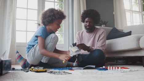 middle aged black father sitting on the floor at home helping his pre teen son to assemble a kit toy, close up, low angle