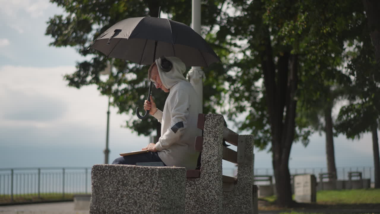 나이 든 백인 남성이 벤치에 앉아 우산 아래에서 헤드폰을 끼고 흐린 날씨의 해안가 풍경을 바라보고 있어요. 나무 그늘과 멀리 보이는 난간과 바다가 보입니다. 은퇴한 작가가 수첩을 확인하며 생각에 잠긴 눈빛으로 느리고 비 오는 분위기 속에서 조용히 앉아 있어요.