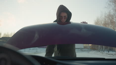 view from inside car shows young man in winter jacket removing support rod and closing bonnet after engine check, with snowy road, bare trees, and cold urban environment visible in background