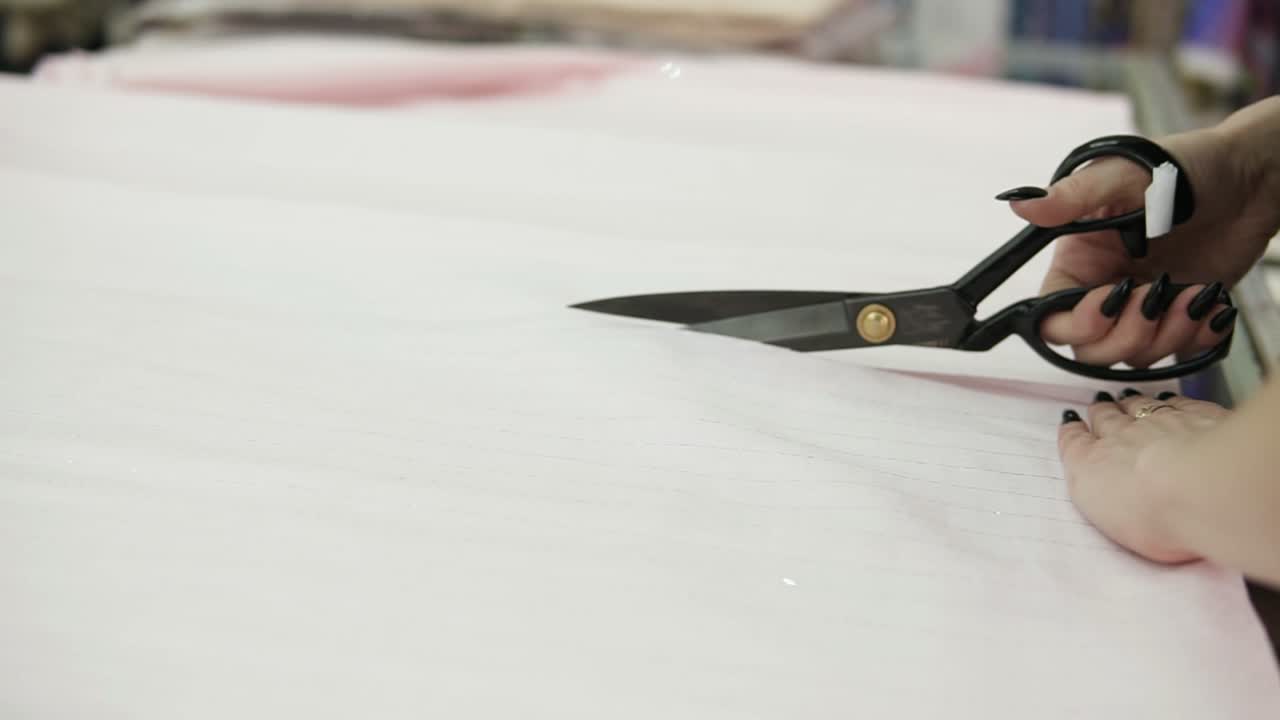 Extremely close up footage of a female seller cutting the light colored tissue using a black scissors. Fabric is on the table