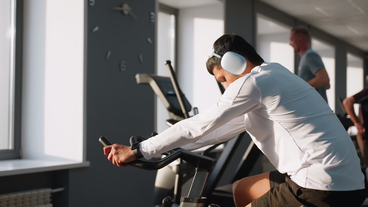 Side view of man in white sportswear listening to music while cycling on stationary bike in bright gym, with another person walking in blurred scene under window light and modern equipment
