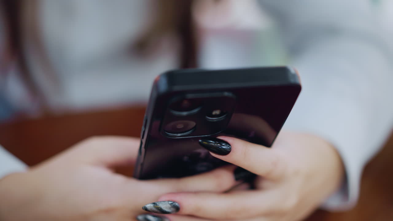 Primo piano di un telefono nero con una donna che digita un messaggio sul telefono con le unghie smaltate, la mano appoggiata su un tavolo di legno marrone, circondato da uno sfondo sfocato.