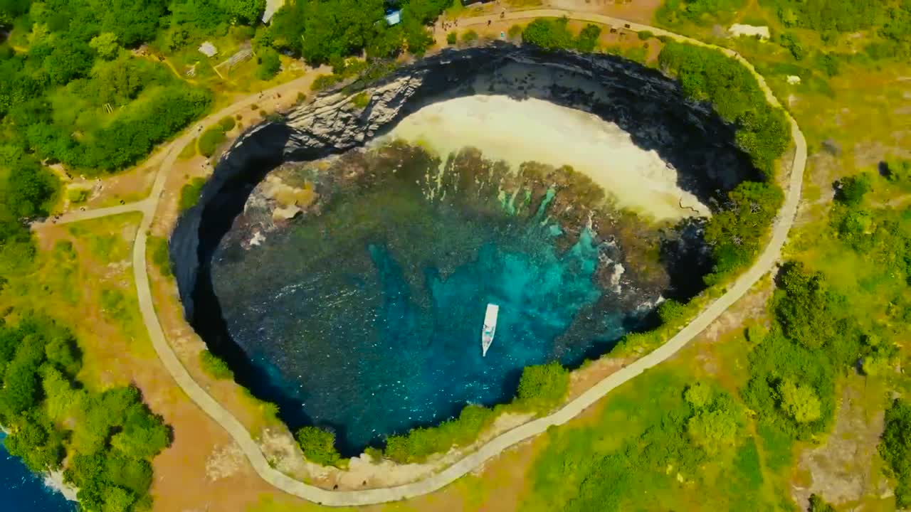 Aerial drone flying higher from Bali Broken beach ground cavity with blue ocean water in it while a large white yacht or ship is in there during a sunny day. Surrounding nature is green with trees.
