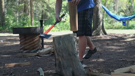 man in t-shirt and shorts splits firewood with axe while camping