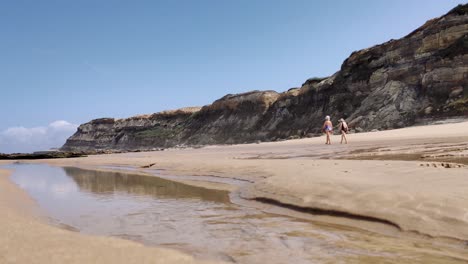 Zwei-ältere-Damen-In-Badebekleidung,-Die-Am-Strand-In-Der-Nähe-Von-Steinklippen-Spazieren,-Wasserstrom-Mit-Niedrigem-Winkel