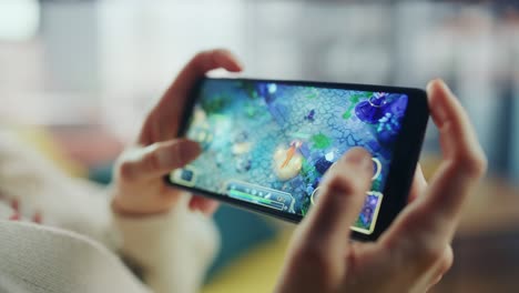 close up on hands playing an rpg strategy video game on a horizontally held smartphone at home living room. feminine hands tapping the mobile phone screen with a colorful game over the internet.