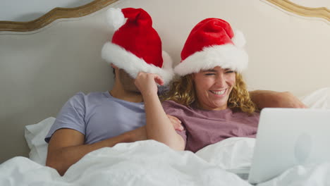 couple at christmas lying in bed at home making video call on laptop wearing santa hats