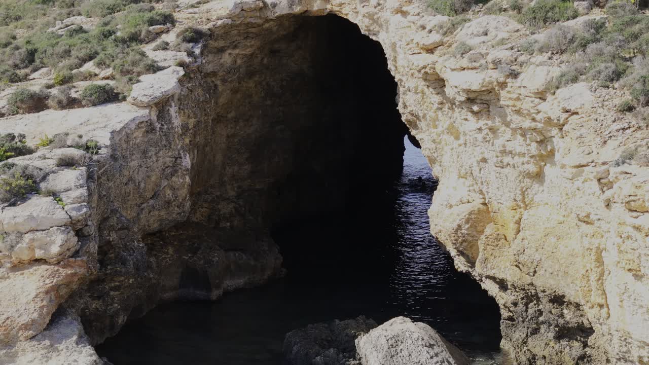 A slow drone pass reveals the Cominotto cave channel at Blue Lagoon, showing a swimmer moving through the sunlit passage between the rocks toward the open sea