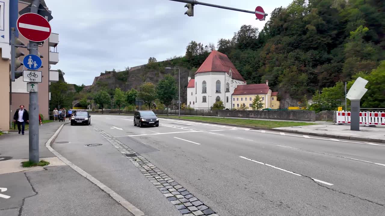Walking across street with view of town houses near river Ilz and church of Saint Salvator on other side of the river bank with daily traffic and street crossing lights in Passau in Germany