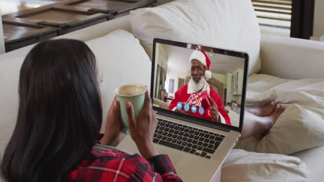mujer biracial en chat de video portátil tomando café durante la navidad en casa