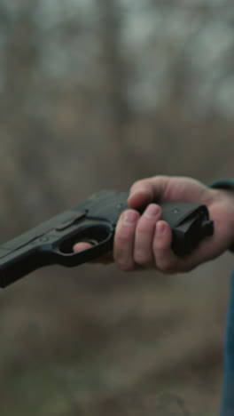 una vista hacia abajo de un hombre con una chaqueta verde y pantalones vaqueros azules sentado en una zona boscosa, profundamente centrado en su teléfono inteligente, con un arma en la mano mirando hacia abajo