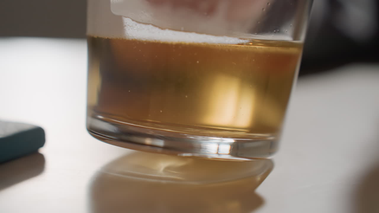 Primo piano di un bicchiere di birra ambrata sul tavolo, mano che inclina il bicchiere lasciando un sottile anello di schiuma, luce calda e soffusa, sottobicchiere riflettente accanto al bicchiere, tranquilla pausa casalinga dopo il lavoro, movimento minimo contemplativo, cinematografico
