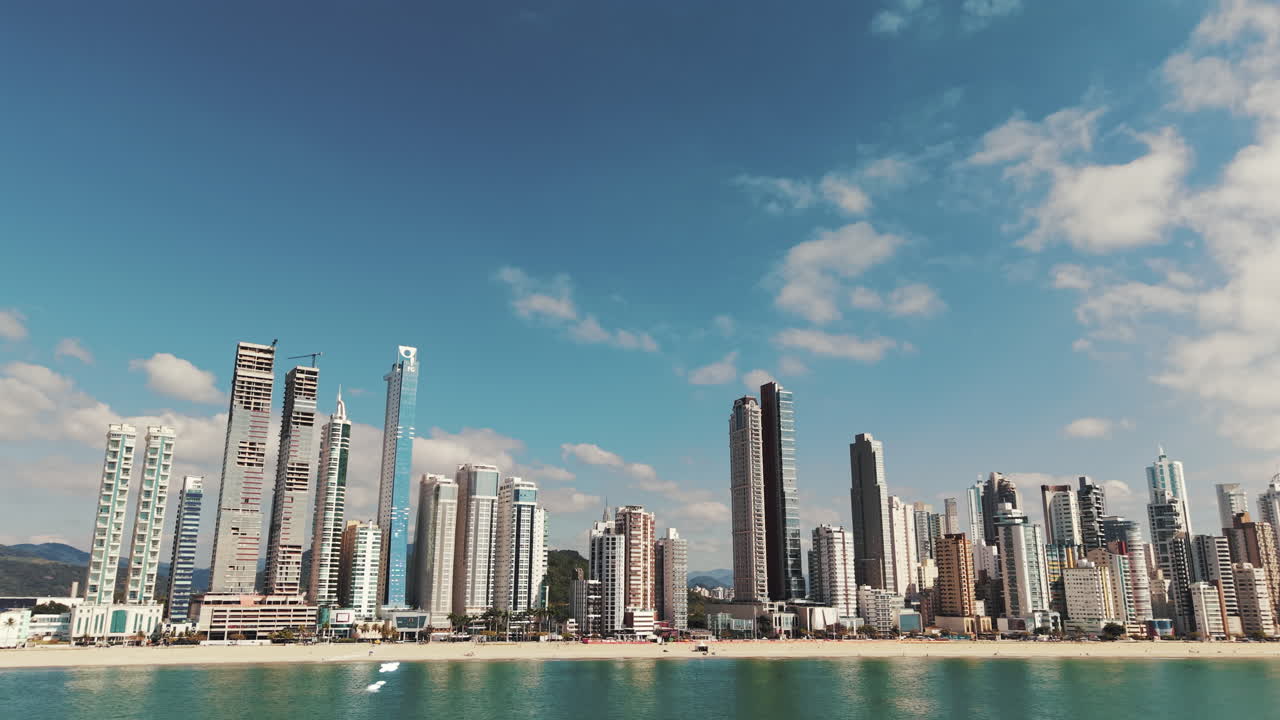 Low-altitude panning shot from the sea showing Balneário Camboriú skyline with tall modern buildings under a blue sky