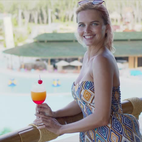 Cheerful-lady-with-fruit-drink-on-balcony-near-swimming-pool