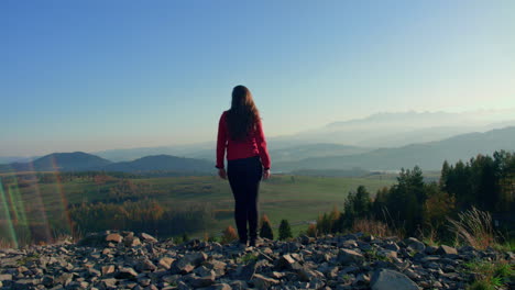 girl walking toward the cliff on a top of the hill looking at landscape filled with hills and mountains