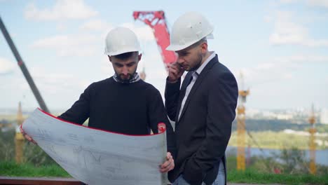 young engineer and foreman with blueprint standing at construction site talking on phone with investor. portrait of two caucasian confident men planning urban infrastructure outdoors on sunny day.