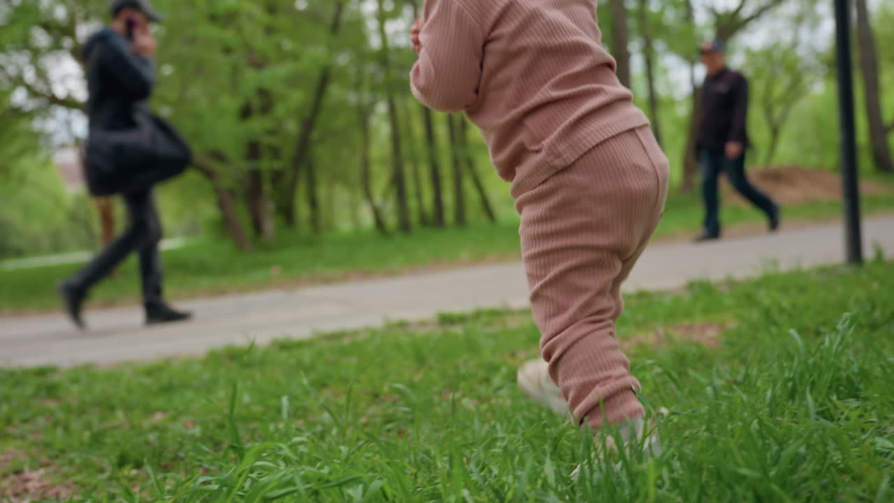 Child Walks Steadily, Little Kid Explores Outdoors Confidently, Young Child Advances Carefully Across Grassy Park, Small Toddler Navigates Through Park With Assistance And Curiosity