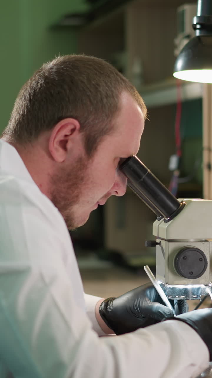 A focused technician in a white lab coat picks something on the table and is working on the circuit under a microscope in a well-equipped laboratory, with various equipment on the desk