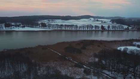 Antena-De-Drones-4k-Uhd-De-Un-Lago-Natural-Rodeado-De-Colinas-Y-Campos-Y-Una-Avenida-De-Robles-En-Baviera,-Alemania,-En-Invierno-Con-Nieve-Durante-Una-Puesta-De-Sol-De-Cielo-De-Nubes-Rojas