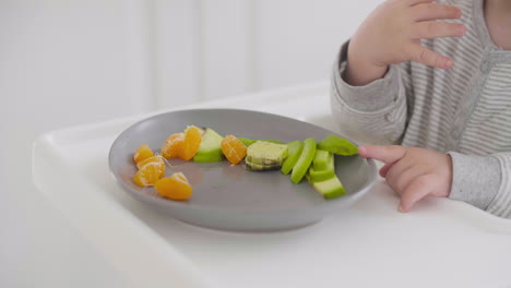 primer plano de un bebé irreconocible tomando segmentos de clementina del plato en una silla alta