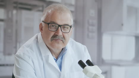 retrato de un médico de laboratorio de cabello blanco con un microscopio mira a la cámara serio y luego sonríe en cámara lenta. imágenes de alta calidad 4k