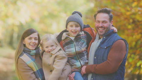 Portrait-Of-Family-With-Mature-Parents-Carrying-Two-Children-In-Front-Of-Autumn-Leaves